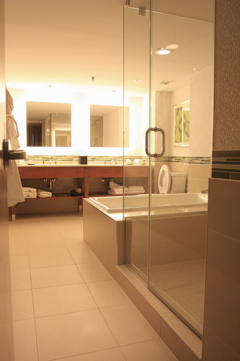 Pale stone bathroom with illuminated mirrors, a vanity and a glass-screened bath area