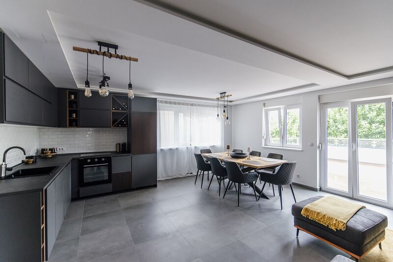 Open-plan room with a run of dark kitchen units, an island and a dining table beside tall windows